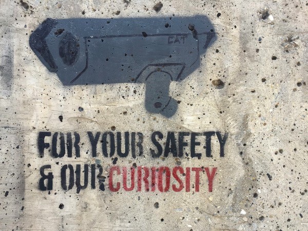 Grafitti de una cámara de seguridad con la frase -For your safety and our curiosity- (Por su seguridad y nuestra curiosidad) escrita debajo